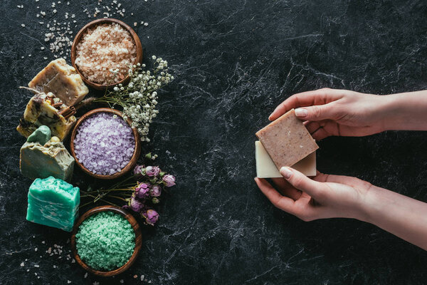 cropped view of female hands with natural homemade soap, dried flowers and sea salt on black marble surface