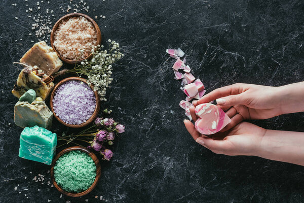 cropped view of female hands with homemade soap in heart shape on black marble surface with dried flowers, natural soap and sea salt for spa