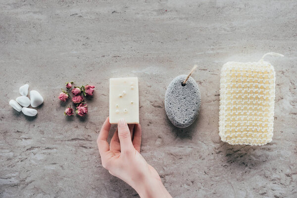cropped view of hand with stones, dried roses, natural soap, pumice and bath sponge for spa on marble surface