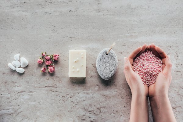 cropped view of hands with sea salt on marble surface with stones, dried roses, natural soap and pumice for spa