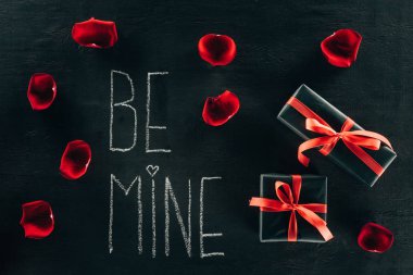 lettering BE MINE surrounded with rose petals and gift boxes on black surface clipart