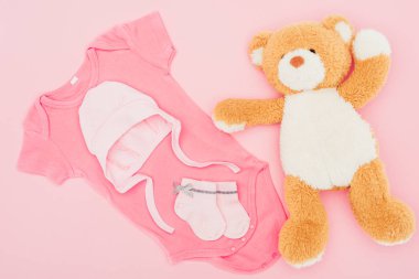 top view of teddy bear with baby clothes isolated on pink clipart