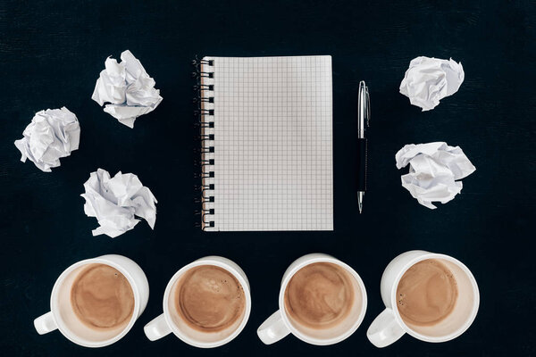 top view of blank notepad with messy crumpled papers and cups of coffee in row isolated on black