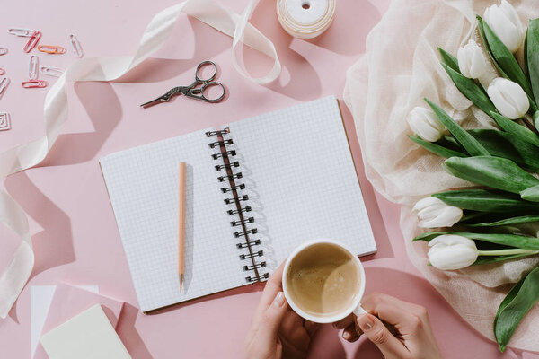 cropped view of woman holding coffee cup on pink surface with tulips and notepad
