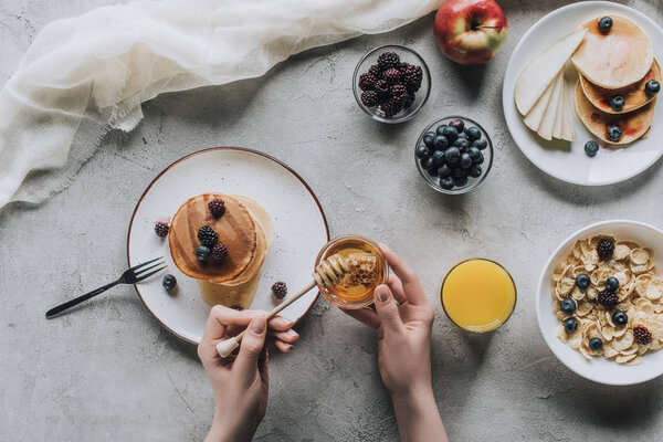 top view of person eating delicious homemade pancakes with honey and fruits on grey