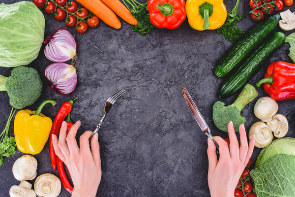 cropped shot of hands holding fork and knife above healthy fresh vegetables on black 