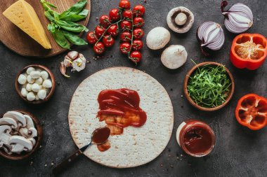 top view of pizza dough with poured ketchup and various vegetables on concrete table clipart