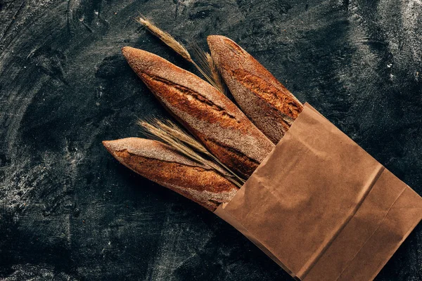 在纸袋和小麦上排列的法国面包的顶部视图在深色桌面上面粉 — 图库照片