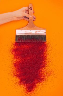 cropped image of woman holding brush above red sand isolated on orange clipart