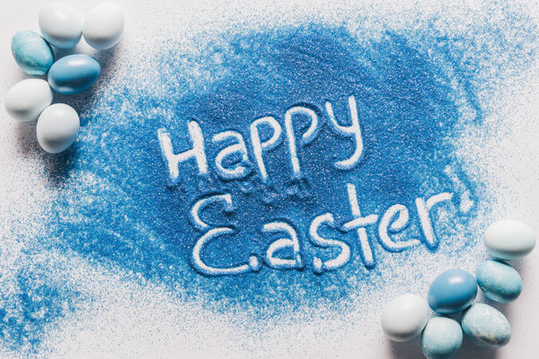 top view of happy easter sign made of blue sand with painted eggs on white