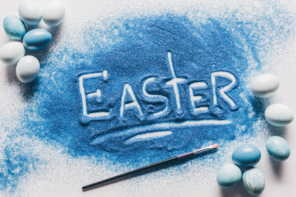 top view of easter sign made of blue sand, brush with painted eggs on white