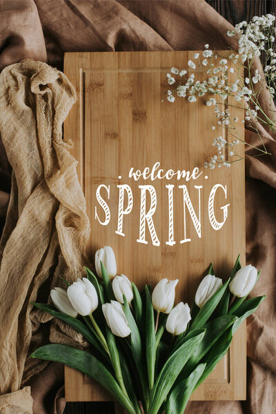 top view of white tulips and WELCOME SPRING lettering on wooden board and tablecloth