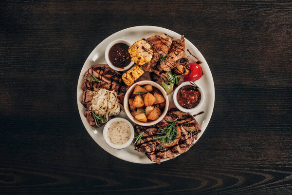 вид сверху на тарелку со стейками из говядины, куриными крылышками и овощами на деревянном столе
