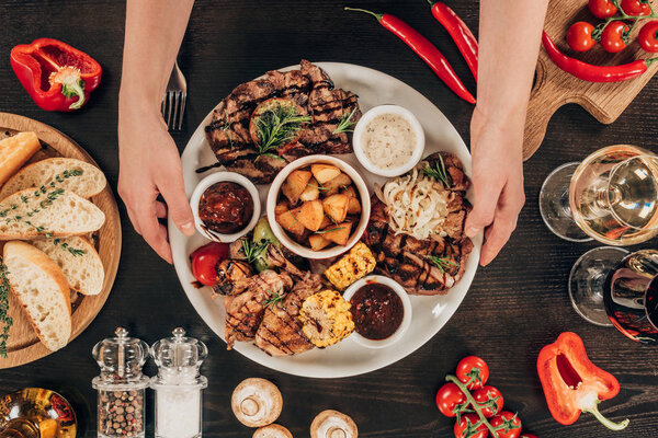 cropped image of woman putting plate with beef steaks, chicken wings and grilled vegetables on table