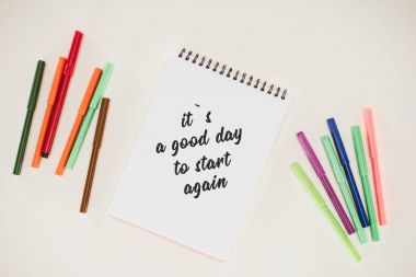 top view of notepad with its a good day to start again lettering and colorful markers isolated on white clipart