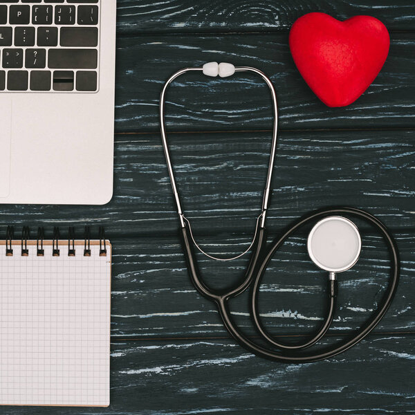 top view of arranged laptop, empty notebook, red heart and stethoscope on dark wooden tabletop