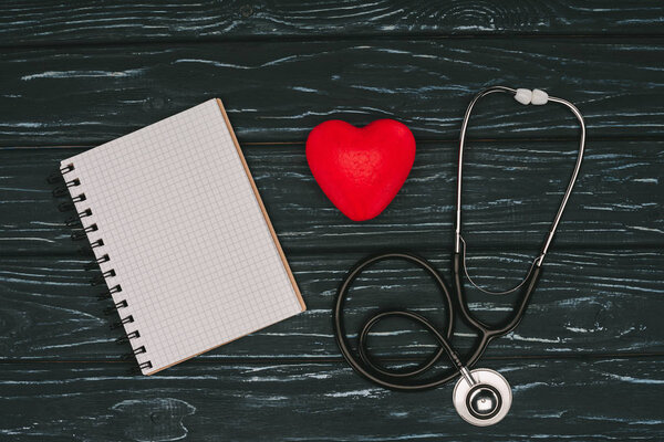 flat lay with arranged red heart, stethoscope and empty notebook on dark wooden tabletop, world health day concept