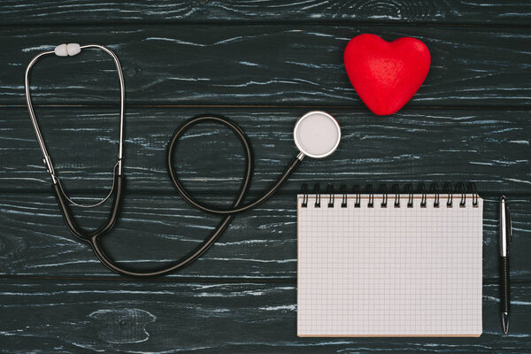 flat lay with arranged red heart, stethoscope and empty notebook on dark wooden tabletop, world health day concept