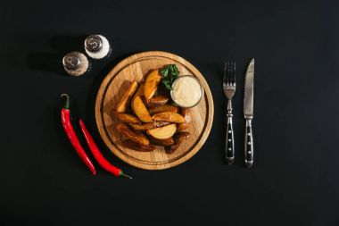 delicious baked potatoes with sauce on wooden board, spices, chili peppers and fork with knife on black   clipart