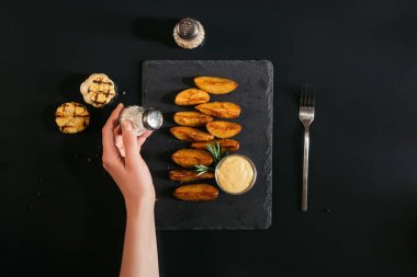 partial top view of person holding salt while eating tasty baked potatoes with sauce on black clipart