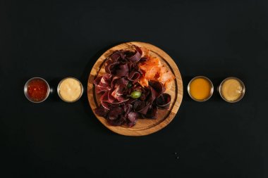 top view of delicious sliced assorted meat on wooden board with various sauces on black clipart