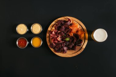 top view of delicious sliced assorted meat on wooden board with glass of beer and sauces on black clipart