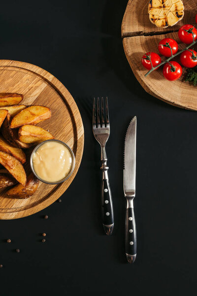 top view of baked potatoes with sauce on wooden board, fork with knife and vegetables on black