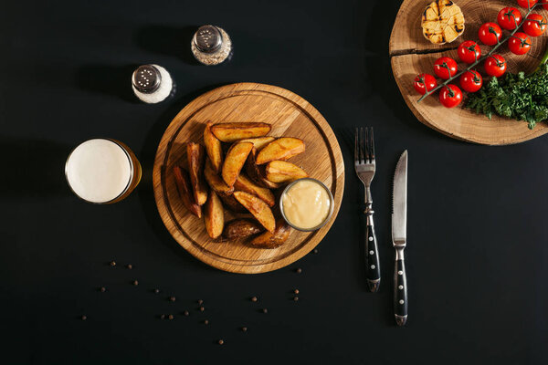 top view of baked potatoes with sauce on wooden board, glass of beer with spices and vegetables on black
