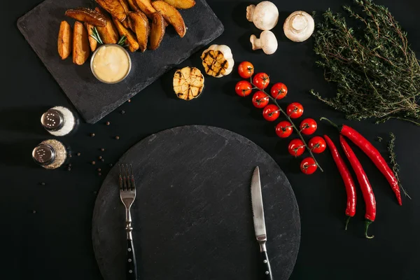 top view of empty slate board with fork and knife, baked potatoes, spices and vegetables on black