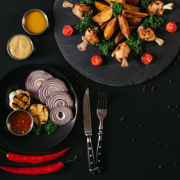 top view of delicious baked potatoes with fried chicken wings, sauces and vegetables on black