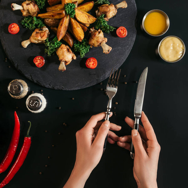 cropped shot of person holding fork and knife while eating delicious roasted potatoes with chicken and vegetables on black