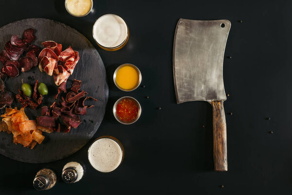 top view of meat knife and gourmet sliced assorted meat on slate board with sauces and spices on black