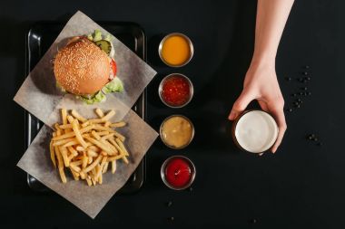 tepsi, çeşitli soslar ve bardak bira siyah üzerinde tutan el Fransız kızartması ile lezzetli burger Üstten Görünüm  