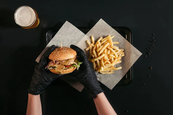 被裁剪的人在手套持有美味的汉堡在托盘上与法式薯条和玻璃啤酒黑色 — 图库照片