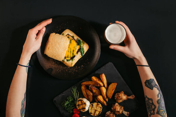 top view of person eating delicious vegan burger with fried potatoes and drinking beer on black