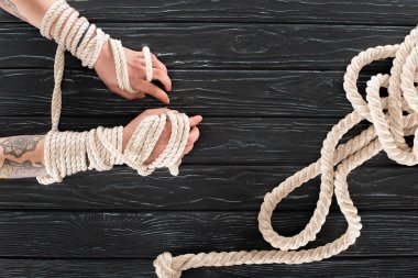 cropped shot of female tattooed hands tied in rope on dark wooden tabletop clipart