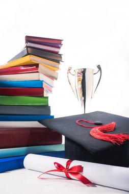 Graduation cap and diploma in front of books and trophy cup clipart