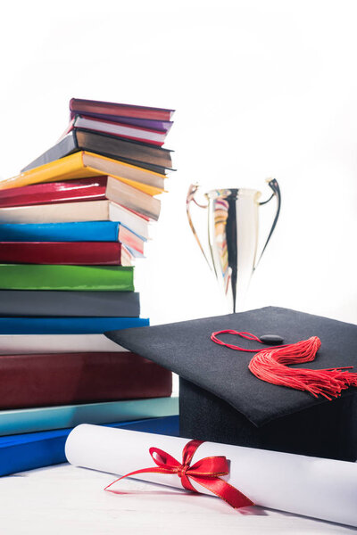Graduation cap and diploma in front of books and trophy cup