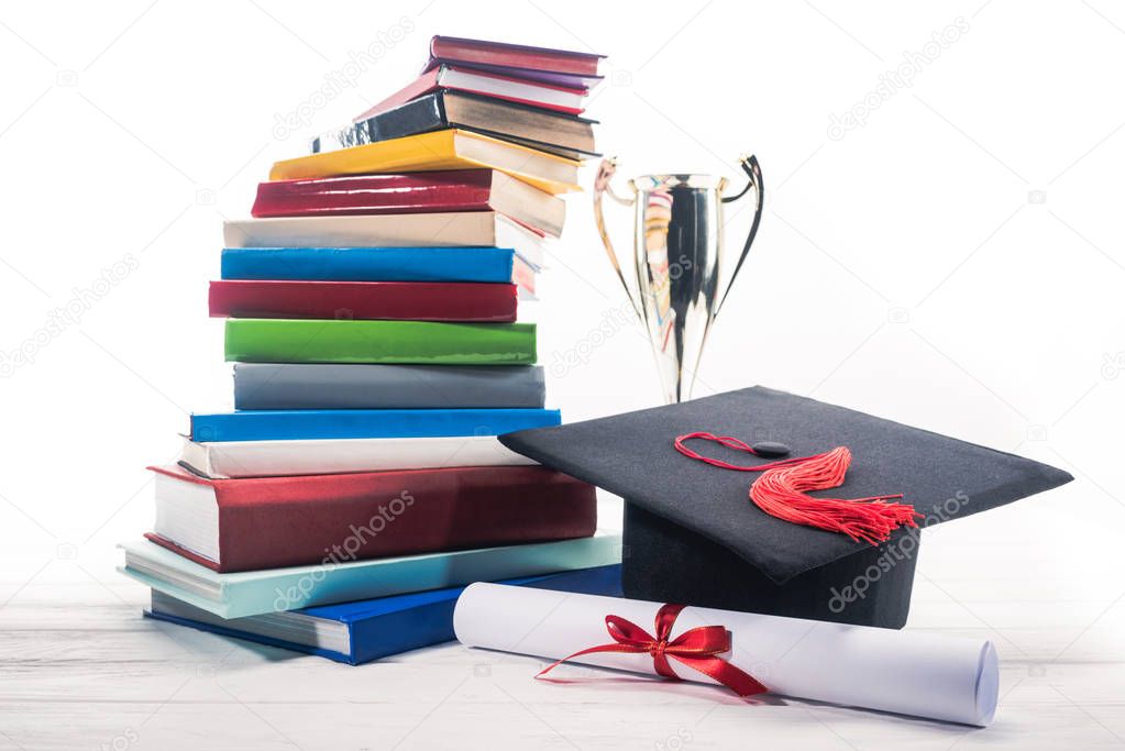 Graduation hat with diploma and trophy cup by stack of books