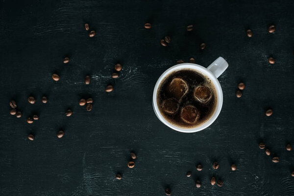 top view of mug of cold iced coffee on dark tabletop with roasted coffee beans around