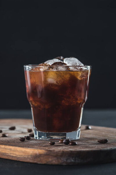 close up view of glass of cold brewed coffee with roasted coffee beans on wooden cutting board on dark backdrop