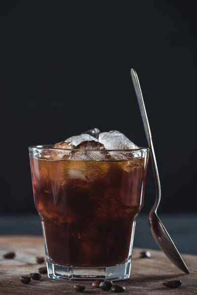close up view of glass of cold brewed coffee with spoon on wooden cutting board on dark backdrop