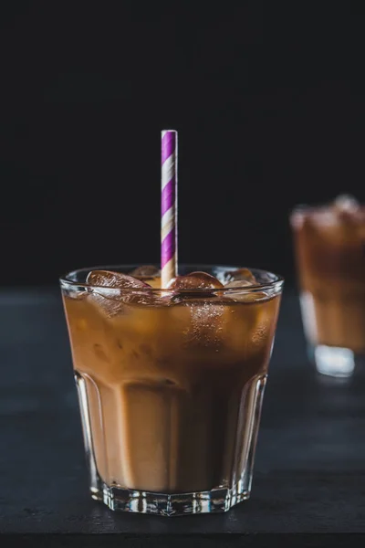 selective focus of glass of cold iced coffee with straw on tabletop on dark backdrop