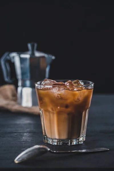 selective focus of glass of cold iced coffee and coffee maker on tabletop on dark backdrop