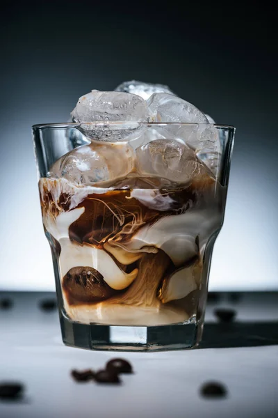 close up view of cold brewed coffee with ice cubes and roasted coffee beans on tabletop in glass on dark backdrop