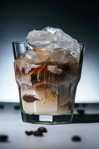 close up view of cold brewed coffee with ice cubes and roasted coffee beans on tabletop in glass on dark backdrop
