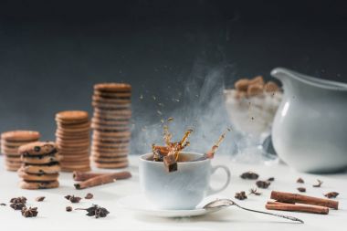 White cup of hot steaming coffee with splashing sugar cubes on table with cookies and spices clipart