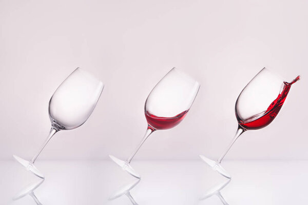 row of inclined wineglasses empty and with splashing wine on reflective surface and on white