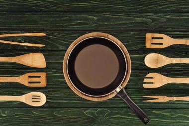 top view of frying pan on round cutting board between wooden kitchen utensils on table  clipart