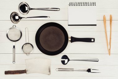 top view of blank textbook, frying pan, butcher axe and kitchen utensils on white wooden table   clipart